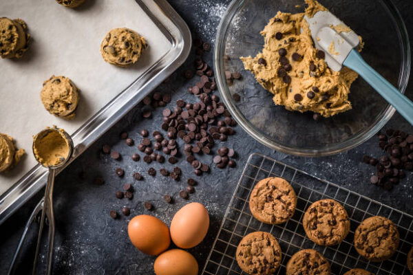 Top view of a crystal bowl full of chocolate chip cookie dough beside a baking tray with a wax paper and cookie dough ready to be baked. Some baked chocolate chip cookies stands on a cooling rack. Predominant colors are brown and bluish grey. Low key DSLR photo taken with Canon EOS 6D Mark II and Canon EF 24-105 mm f/4L